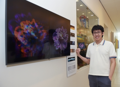 Ethan Tseng standing in the hallway of the Friend Center next to a large TV monitor displaying 2 images of flowers side by side. The flower on the right is a clearer image. Ethan is holding a small postcard of the clearer flower.