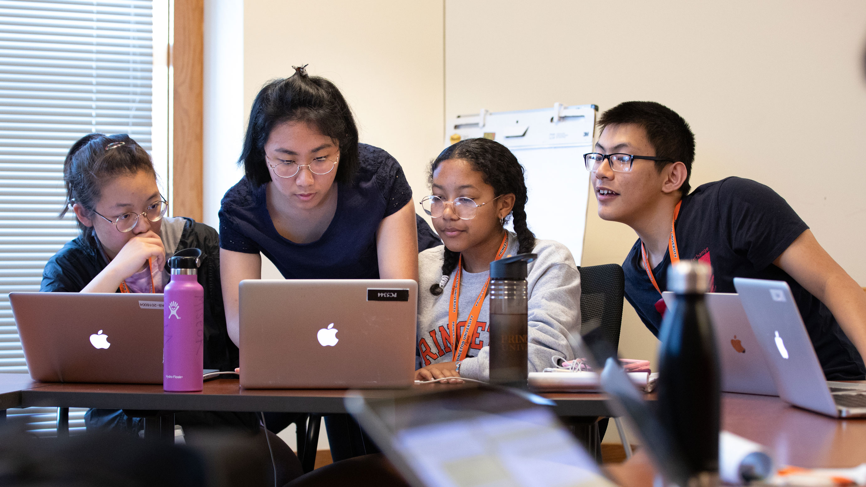 Four high school age students sitting in a classroom looking at the same laptop.